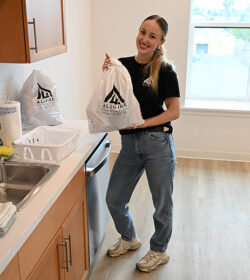 A woman standing in a kitchen holding a bag of supplies with the Allgire Foundation logo on it